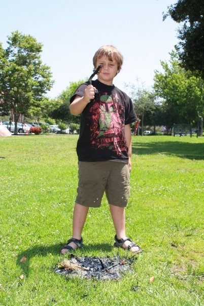 A boy looking at a stick with tar on it while standing next to a small pool of asphalt in the grass.